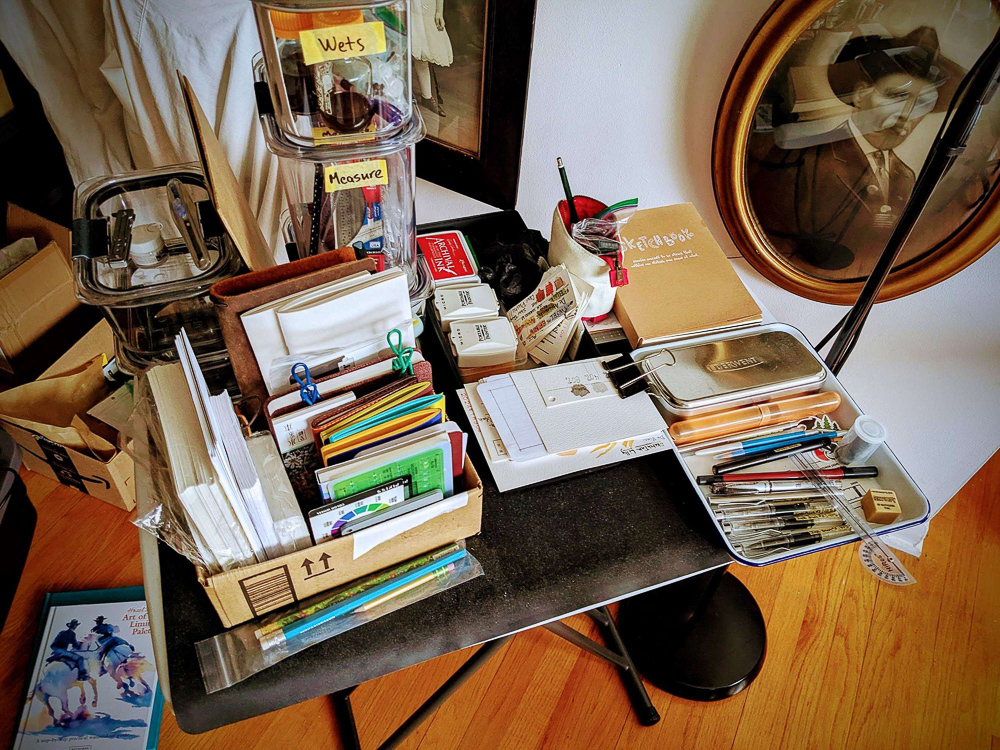 A small table full of art supplies including: Too many fountain pens and many incomplete pocket sketchbooks